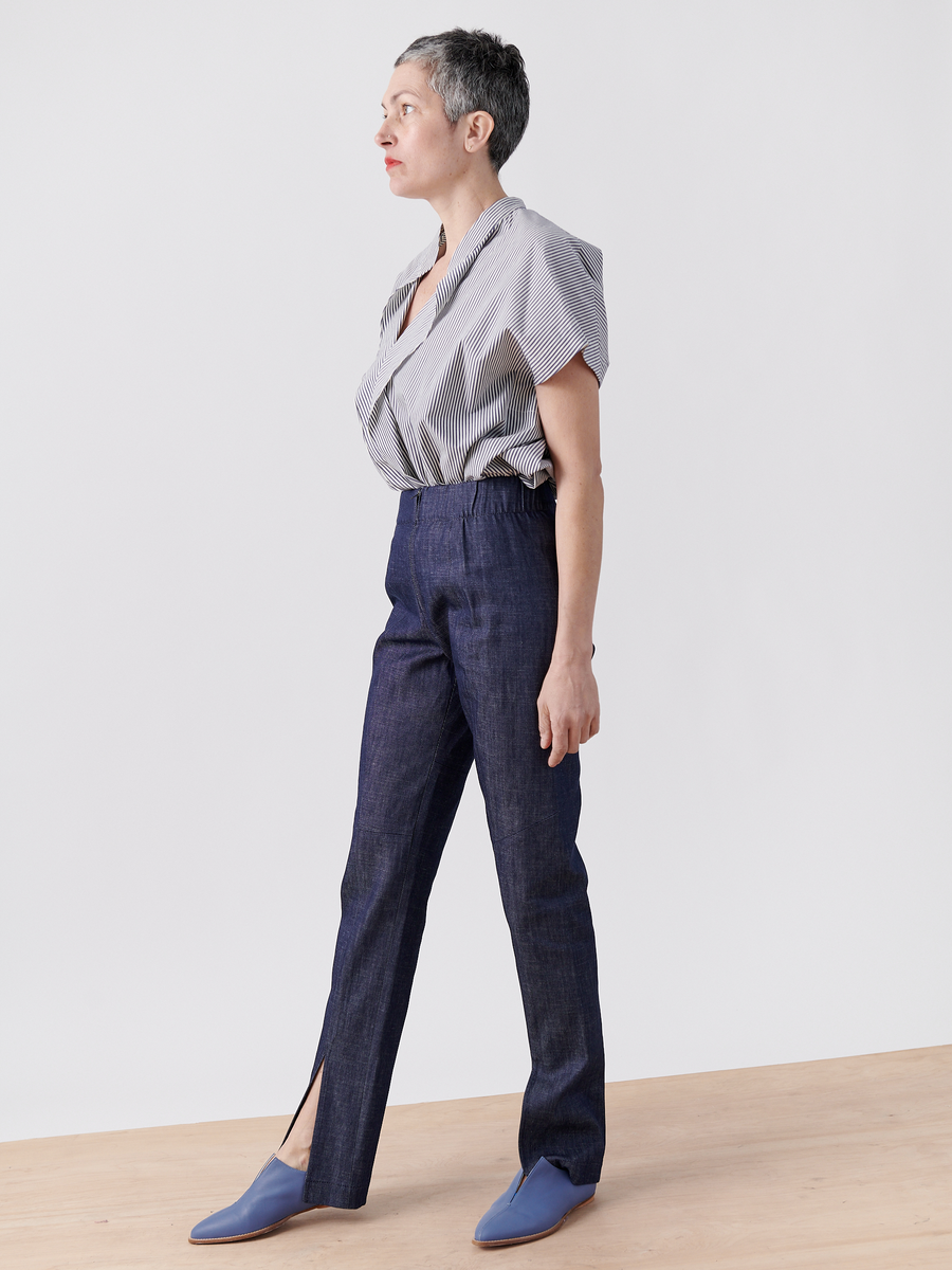 A person with short gray hair stands on a wooden floor against a light background, wearing a gray-striped blouse and Zero + Maria Cornejo's Denim Tala Trouser - Final Sale, dark blue with ankle slits. Their look is completed by blue shoes, and they gaze off to the side neutrally.