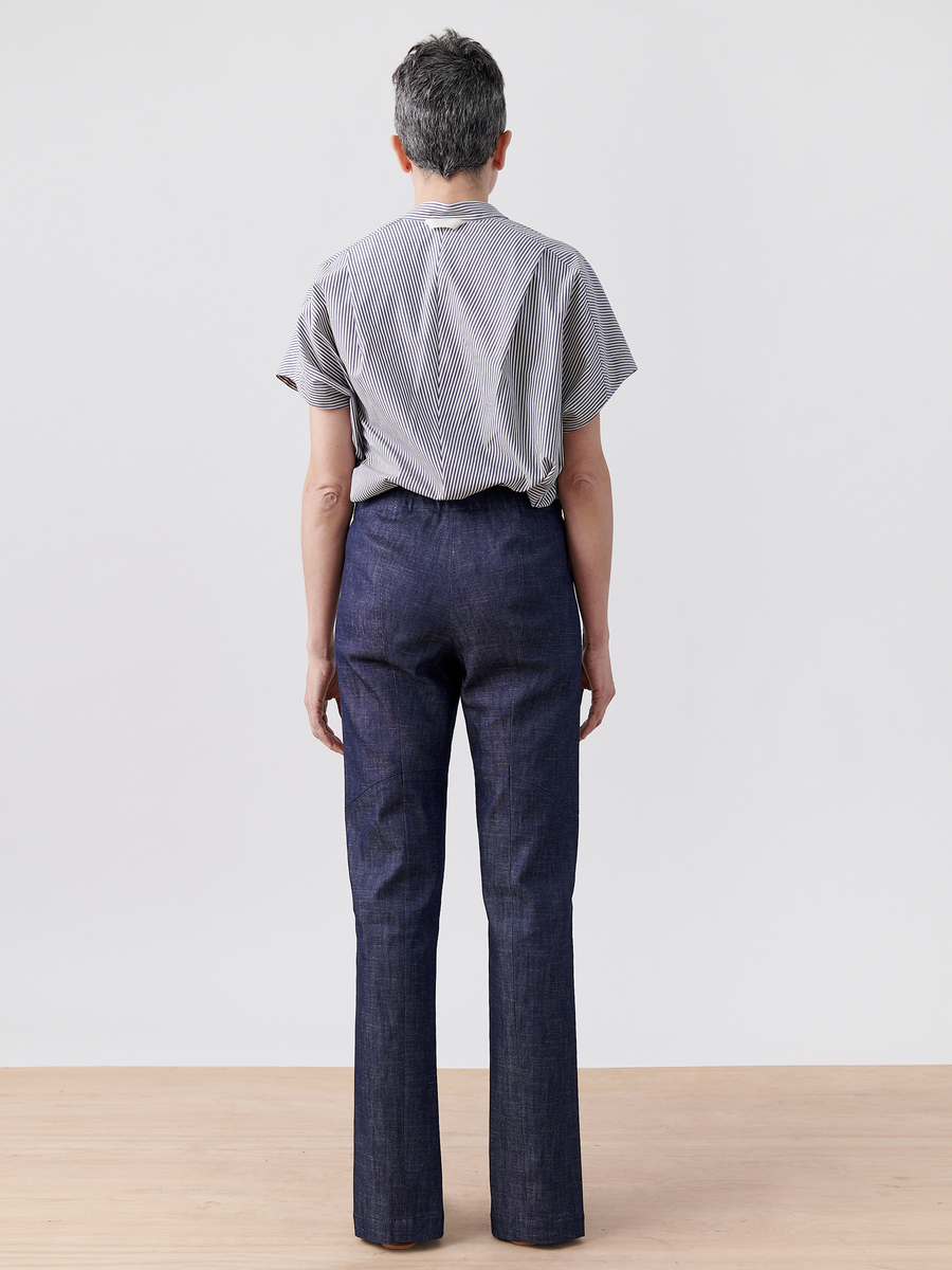 A person with short gray hair faces away, wearing a striped short-sleeve shirt and organic Denim Tala Trousers by Zero + Maria Cornejo. They stand on a wooden surface against a plain light gray background.