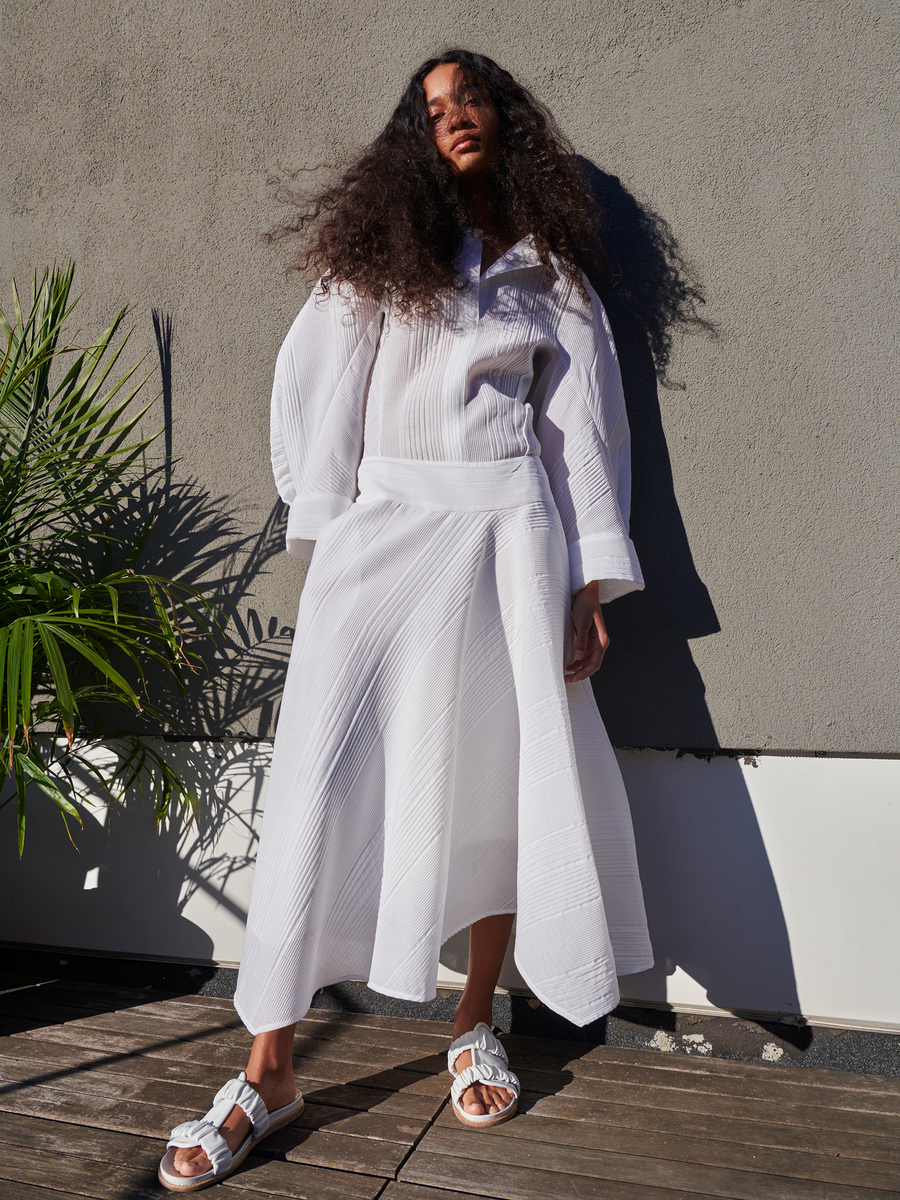 A person with long, curly hair stands on a wooden deck in a white two-piece outfit that includes the Aki Wave Skirt by Zero + Maria Cornejo. The asymmetrical skirt and white sandals contrast with the gray wall and lush green plant, highlighted by sunlight creating distinct shadows.