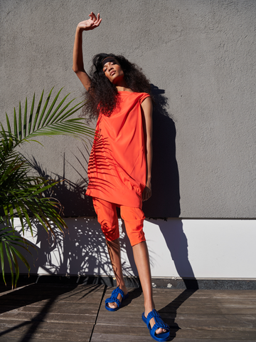 A person in orange silk charmeuse Crop Curve Ori Pant - Final Sale by Zero + Maria Cornejo stands against a gray wall, partially shaded. Curly hair frames their face as they raise an arm, paired with blue sandals near a potted plant on a wooden deck.